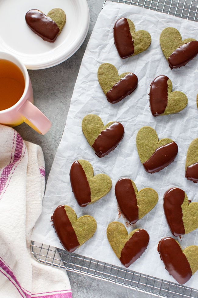 Matcha Cookies from thelittlekitchen.net Chocolate Dipped Heart Shaped Matcha Cookies on parchment paper and cooling rack with kitchen towel, pink cup of tea, and cookie on plate in the background from thelittlekitchen.net