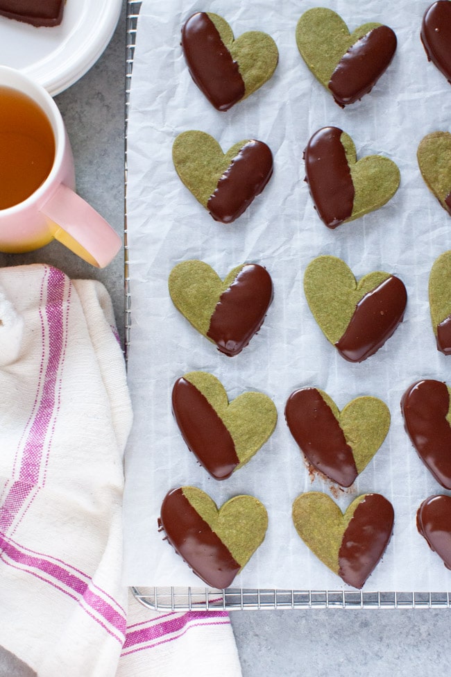Matcha Cookies from thelittlekitchen.net Heart Shaped Chocolate Dipped Matcha Cookies on white parchment paper, next to kitchen towel, cup of tea, and white plate from thelittlekitchen.net