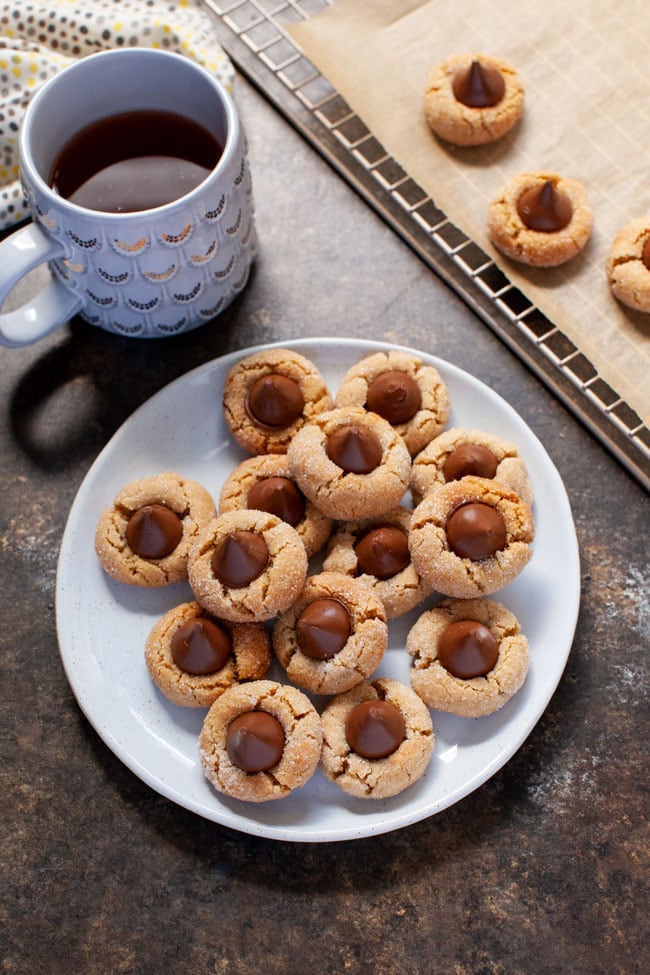 Peanut Butter Blossom Cookies from thelittlekitchen.net Peanut Butter Cookies topped with a Hershey Kiss sit on a off-white plate next to a cup of coffee.